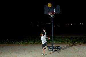 A child shoots a basketball at a hoop during nighttime practice in an outdoor playground with city lights in the background photo