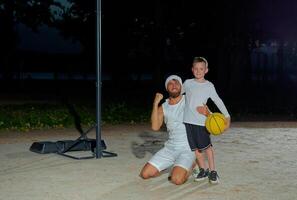 Positive boy holding in hand basketball ball and hugging handsome dad smiling to camera standing outside in open air court. photo