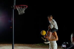 A father joyfully lifts his son for a basketball shot under the night sky at a local court photo