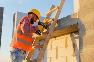 Construction worker using cordless drill on wooden ladder at building site photo