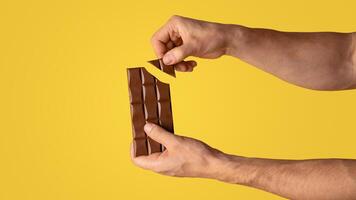 Closeup view of man breaking piece of chocolate from bar, orange background. Copy space. Panorama photo