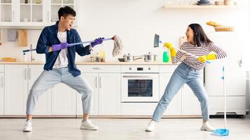 Crazy asian young couple having fun while cleaning apartment, joyful chinese man and woman with brush and mop imitating fight while house-keeping, kitchen interior, full length, copy space photo
