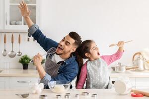 Cheerful arab father and little daughter having fun while baking together in kitchen, happy middle eastern dad and child singing and fooling, using spatula and whisk as microphones, copy space photo