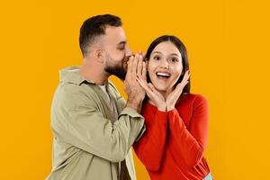 A young couple shares an affectionate moment with the man whispering in the woman's ear. They are smiling and enjoying each other's company against a vibrant yellow backdrop. photo