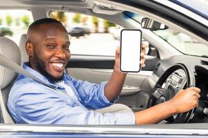 Cheerful young black man sitting in luxury car, showing cellphone with white blank screen, recommending newest mobile application for navigation, mockup. Automobile, driving and modern technologies photo