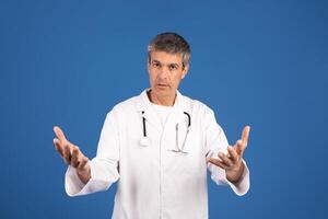 A mature Caucasian man dressed in a white doctor's coat expresses engagement and concern, using open hand gestures to communicate. The plain blue background emphasizes his presence. photo