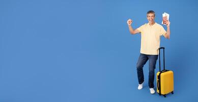A cheerful middle aged man stands against a blue background, holding up his passport and displaying excitement for his upcoming travels. He has a suitcase beside him, ready for adventure, copy space photo
