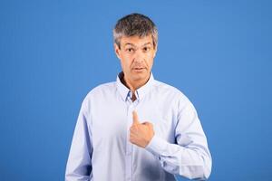 A middle aged Caucasian man stands against a blue background, pointing to himself with an expression that suggests self-reflection or introduction. His calm demeanor conveys confidence. photo