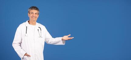 A middle aged European man dressed in a white lab coat stands isolated against a blue backdrop, smiling and inviting, copy space photo