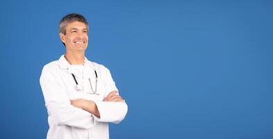 A mature European man dressed in a white doctor's coat stands confidently with arms crossed. He has a friendly smile, conveying professionalism and warmth in a healthcare setting, copy space photo