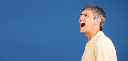 A middle aged man in his 50s yells out in frustration while standing alone against a vibrant blue background, conveying a sense of isolation and emotional intensity, copy space photo