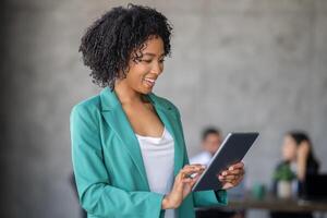 A woman smiles while interacting with a tablet in a contemporary office setting. Background features a diverse team engaged in discussions, showcasing teamwork and innovation in business. photo