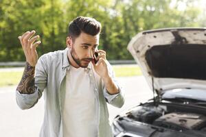 Upset young Caucasian man standing near his broken car with open hood, experiencing engine failure, having mobile phone conversation, calling breakdown road service on cellphone photo
