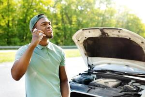 Stressed black guy standing near broken vehice, having mobile phone conversation with breakdown road service, asking for assistance in car wreck. African American man calling emergency help number photo