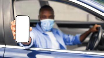 Closeup of black man in surgical mask showing mobile phone with white empty screen for mock up sitting on driver seat in car, holding gadget in hand, selective focus on device, covid protection photo