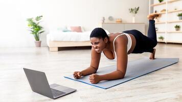 Plus size Afro lady working out at home with online personal trainer, using laptop, standing in elbow plank pose. Overeight black woman training to tutorial on web, indoors photo