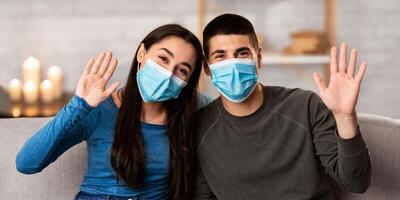 Smiling couple wearing surgical face mask sitting on sofa at home and waving hands to camera, showing hello sign photo