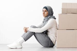 Upset arab family sitting separated by boxes on floor in empty apartment, having problem with relocating, white wall background, side view, copy space. Depressed middle-eastern couple at new home photo