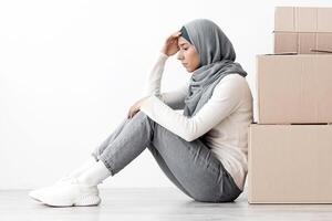 Frustrated arab couple sitting by paper boxes on floor in empty apartment, having problem with relocating, white wall background, side view, copy space. Depressed middle-eastern family at new home photo