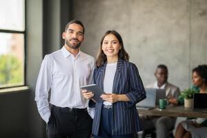 Two professionals stand together in a stylish office, engaging with digital devices. Behind them, a diverse team discusses projects, reflecting teamwork and international cooperation. photo