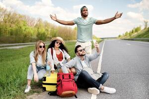 Unhappy millennial tourists sitting on roadside, cannot catch car, waiting for too long, having no smartphone connection. Young friends hitchhiking on highway, having problem finding free ride photo