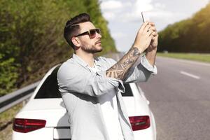 Upset millennial man standing on road next to broken auto, raising hand with cellphone, trying to call roadside assistance. No connection, car breakdown, bad mobile network concept photo