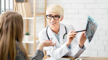 Consultation with doctor in clinic. Adult female therapist in coat and glasses shows x-ray and explains problem to patient in modern office interior at table with tablet and tonometer, free space photo