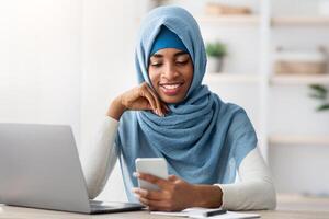Black Islamic Business Lady In Hijab Using Smartphone While Working At Desk In Office, Young African Muslim Woman Browsing New App On Mobile Phone, Messasing Or Shopping Online, Copy Space photo