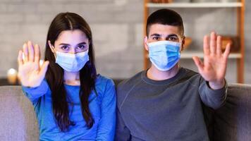 Stop Coronavirus Outbreak. Young couple in protective surgical masks gesturing stop sitting on the couch at home with pc photo