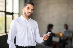 A confident man in a white shirt holds a smartphone while standing in a corporate office. In the background, a diverse team collaborates on a project, reflecting active teamwork. photo