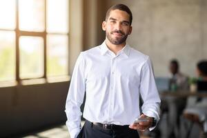 A confident man in a white shirt holds a smartphone while interacting with his diverse team in a corporate office. The atmosphere is focused on teamwork and achieving success during their meeting. photo