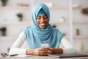 Portrait Of Smiling African Islamic Lady In Hijab Using Smartphone In Office, Cheerful Black Muslim Woman Sitting At Desk With Cellphone In Hands, Browsing Internet, Shopping Online Or Messaging photo