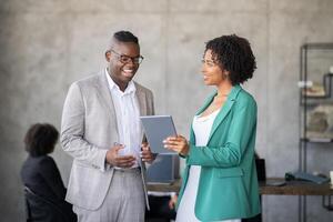Two professionals engage in a discussion while reviewing a digital tablet in a contemporary office. They showcase teamwork and cooperation in a diverse business setting. photo