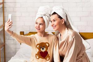 Fun spa day. Beautiful mother with her daughter in bath towels taking selfie on bed at home photo