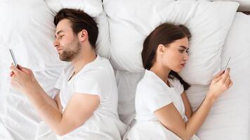 Marital Crisis, Indifference. Couple Using Cellphones Lying Back-To-Back In Bed Indifferent To Each Other. Millennial Spouses Ignoring One Another Texting With Lovers At Home. View From Above photo