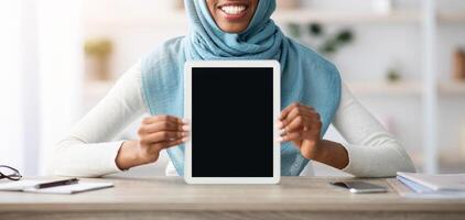 Smiling Black Muslim Lady In Hijab Demonstrating Digital Tablet With Blank Screen While Sitting At Desk In Home Office, African Islamic Female Showing Copy Space For Your Website Or App Design photo