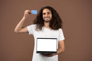 A young man with long curly hair smiles while holding a laptop and a credit card. The setting is simple and isolated, emphasizing his relaxed lifestyle and connection to technology. photo