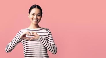 Cheerful young asian lady showing heart gesture with hands near chest, smiling korean woman making love sign and looking at camera, kind millennial female standing on pink background, copy space photo