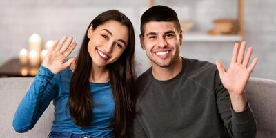 Hello. Smiling couple sitting on sofa at home and waving hands to camera, showing hi sign photo