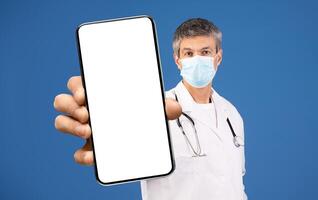 A middle aged Caucasian man, dressed in a white lab coat and wearing a medical mask, is holding a blank smartphone. He appears focused, representing modern healthcare in a contemporary setting. photo