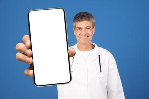 A mature Caucasian doctor in a white coat holds a blank smartphone in front of him. He is smiling, posing against a blue background, promoting digital health solutions. photo