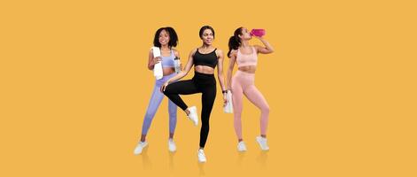 Three women of diverse backgrounds display fitness enthusiasm by stretching, posing, and hydrating in a vibrant setting. The bright yellow background adds energy and motivation to their workout. photo