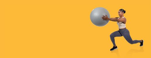 A woman engages in a lunge exercise while holding a stability ball, demonstrating strength and balance. The bright yellow background adds energy to her workout routine. photo