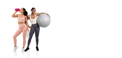 Two women perform fitness activities in a modern studio. One holds a water bottle while the other leans against an exercise ball. They showcase an active lifestyle and motivation. photo