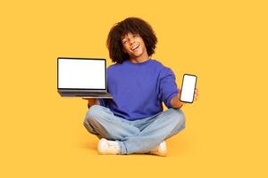 A lively young man with curly hair sits cross-legged against a vibrant yellow backdrop. He holds a laptop in one hand and a smartphone in the other, radiating a sense of energy and innovation. photo