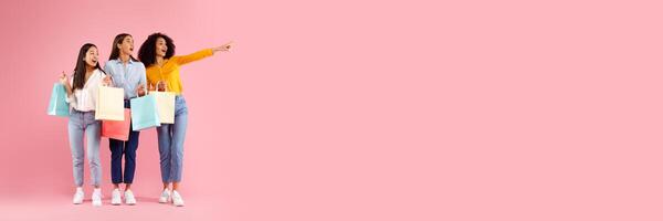 Three excited diverse ladies with colorful shopping bags pointing and looking aside, standing over pink studio background, panorama with copy space, full length photo