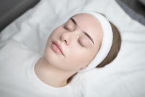 A young woman lies comfortably on a treatment table in a beauty salon. She is undergoing a long-lasting makeup procedure, with soft lighting creating a serene atmosphere for pampering. photo