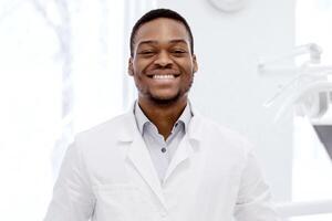 Portrait Of Handsome Young Black Dentist Doctor In Uniform Posing In Clinic. Professional Affican American Orthodont Standing In Modern Stomatological Cabinet And Smiling At Camera, Free Space photo