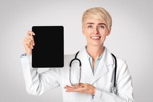 Device for work with health protection in clinic. Smiling adult lady in white coat shows with hand on tablet with empty screen with modern medical app, isolated on gray background, studio shot photo