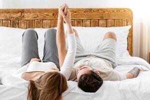 Happy relationship and free time at home during quarantine and self-isolation. Young husband and wife hold hands, lie on wooden bed in modern interior of cozy country bedroom, vertical, free space photo